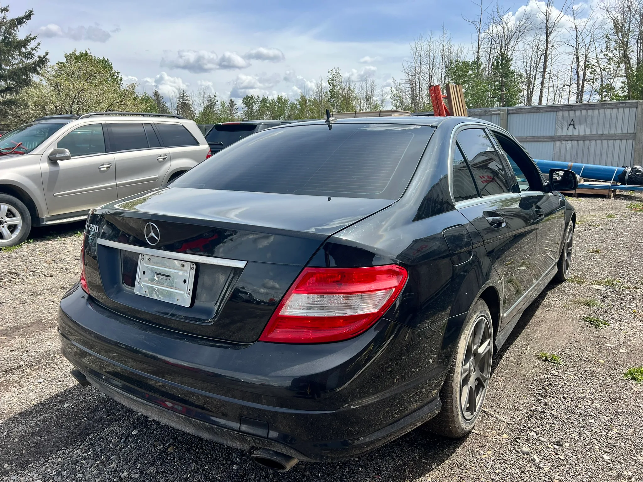 Black Mercedes-Benz sedan with tinted windows and dual exhaust parked on gravel lot; dealer plate visible, metal fence and trees in background.