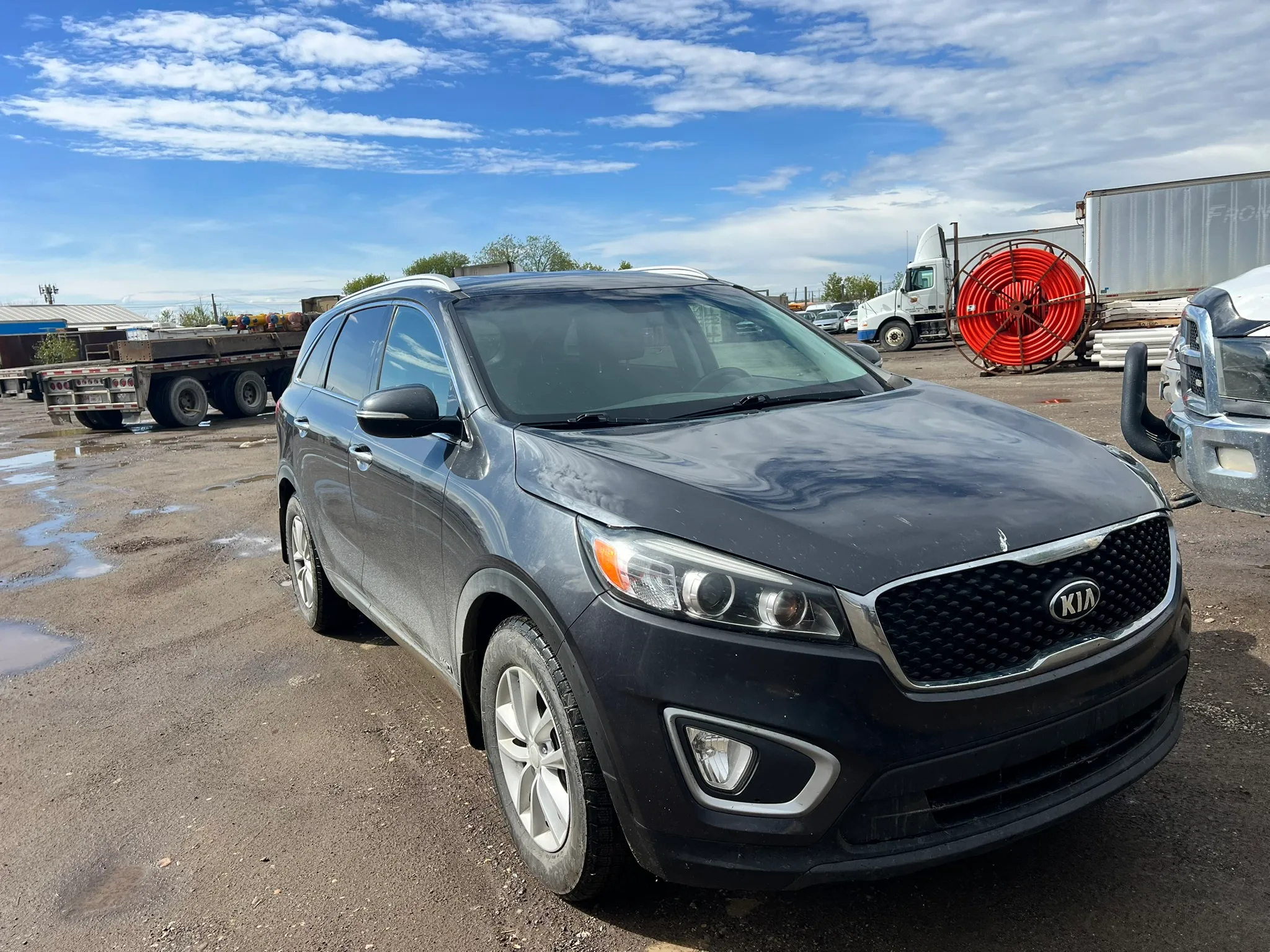 Black Kia Sorento SUV parked on gravel lot with clean bodywork and visible Kia grille; industrial equipment and semi-trailer in background.
