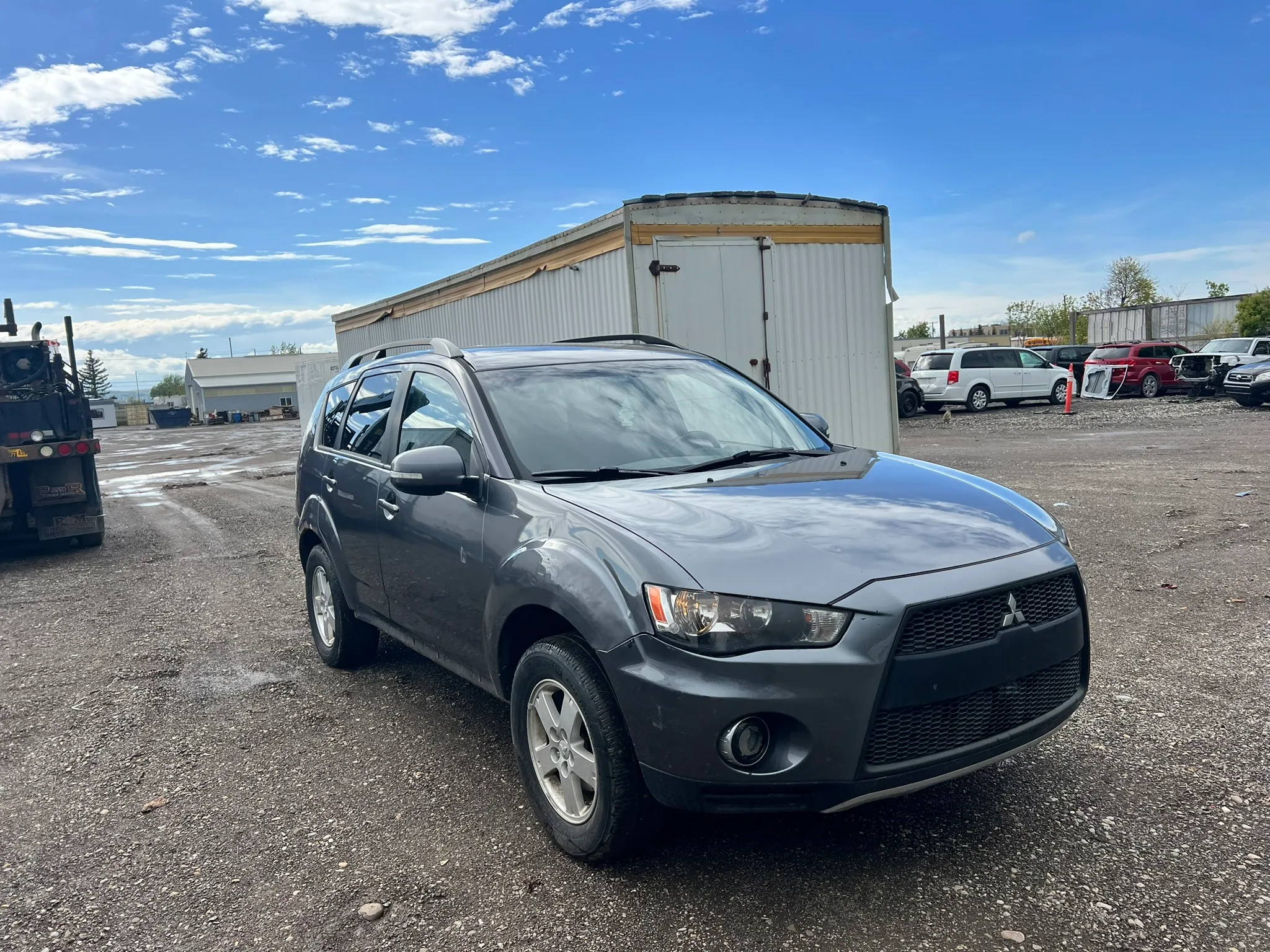 Gray Mitsubishi Outlander angled on gravel lot with clean body and intact headlights; white metal building and scattered clouds in background.
