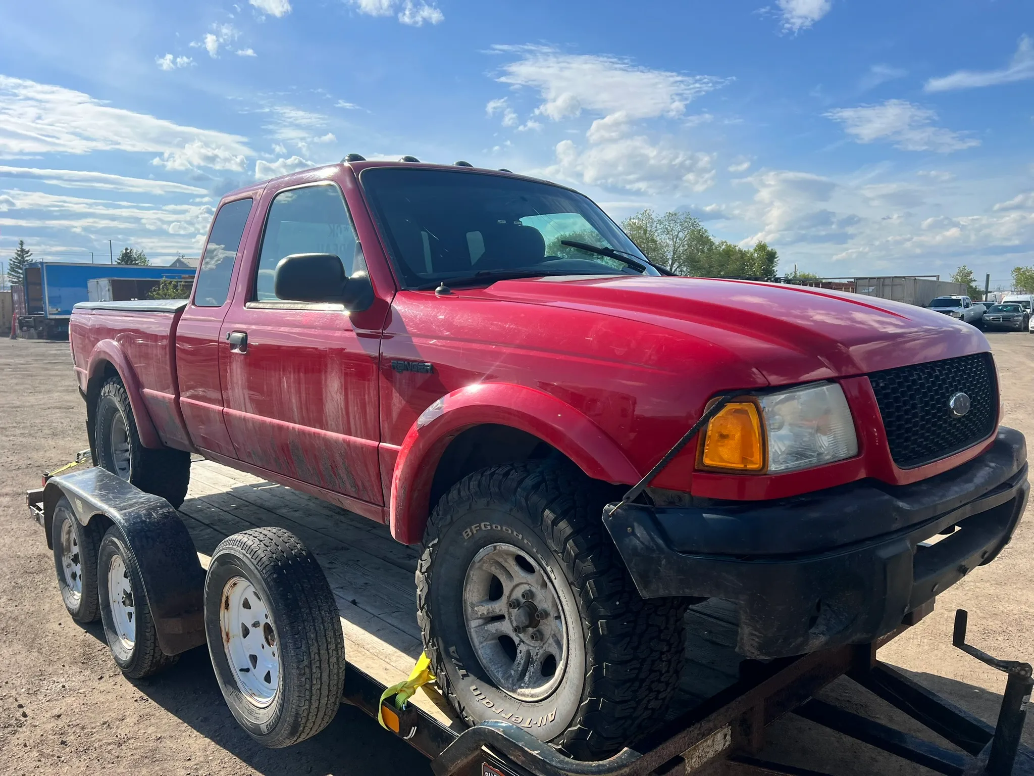Red Ford Ranger with extended cab and off-road tires secured on dual-axle flatbed trailer; dirt lot and trees in background.