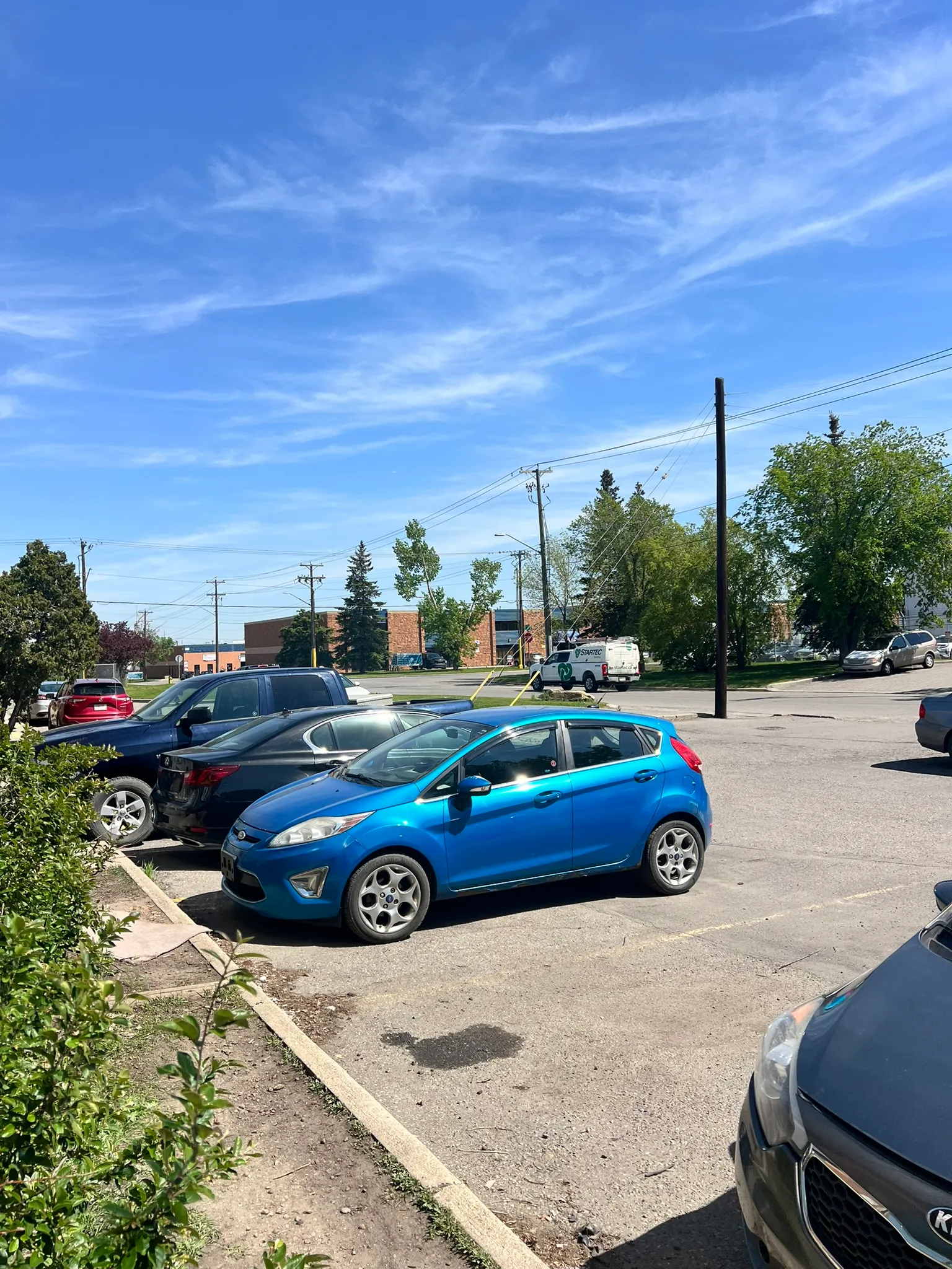 Bright blue compact car parked in sunlit lot with black and silver vehicles nearby; utility poles, trees, and buildings under clear sky with scattered clouds.
