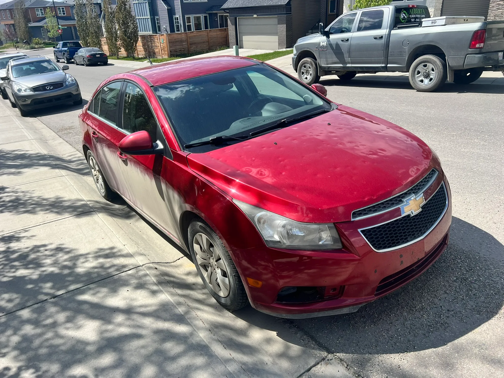 Red Chevrolet Cruze parked close to curb on residential street; dusty exterior, modern houses and other vehicles in background.