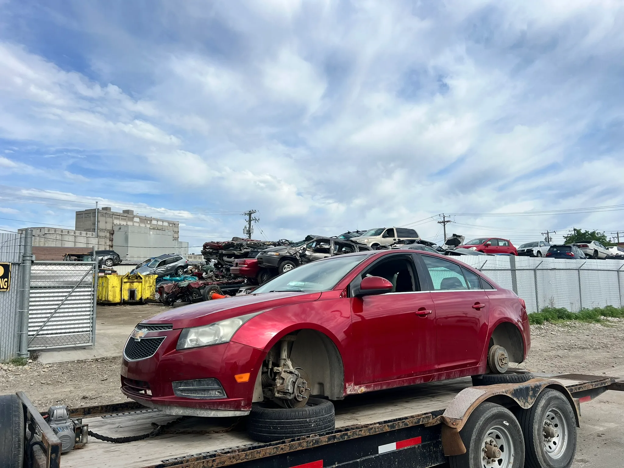 Red Chevrolet Cruze missing front wheels secured on flatbed trailer; junkyard with stacked vehicles and chain-link fence in background.