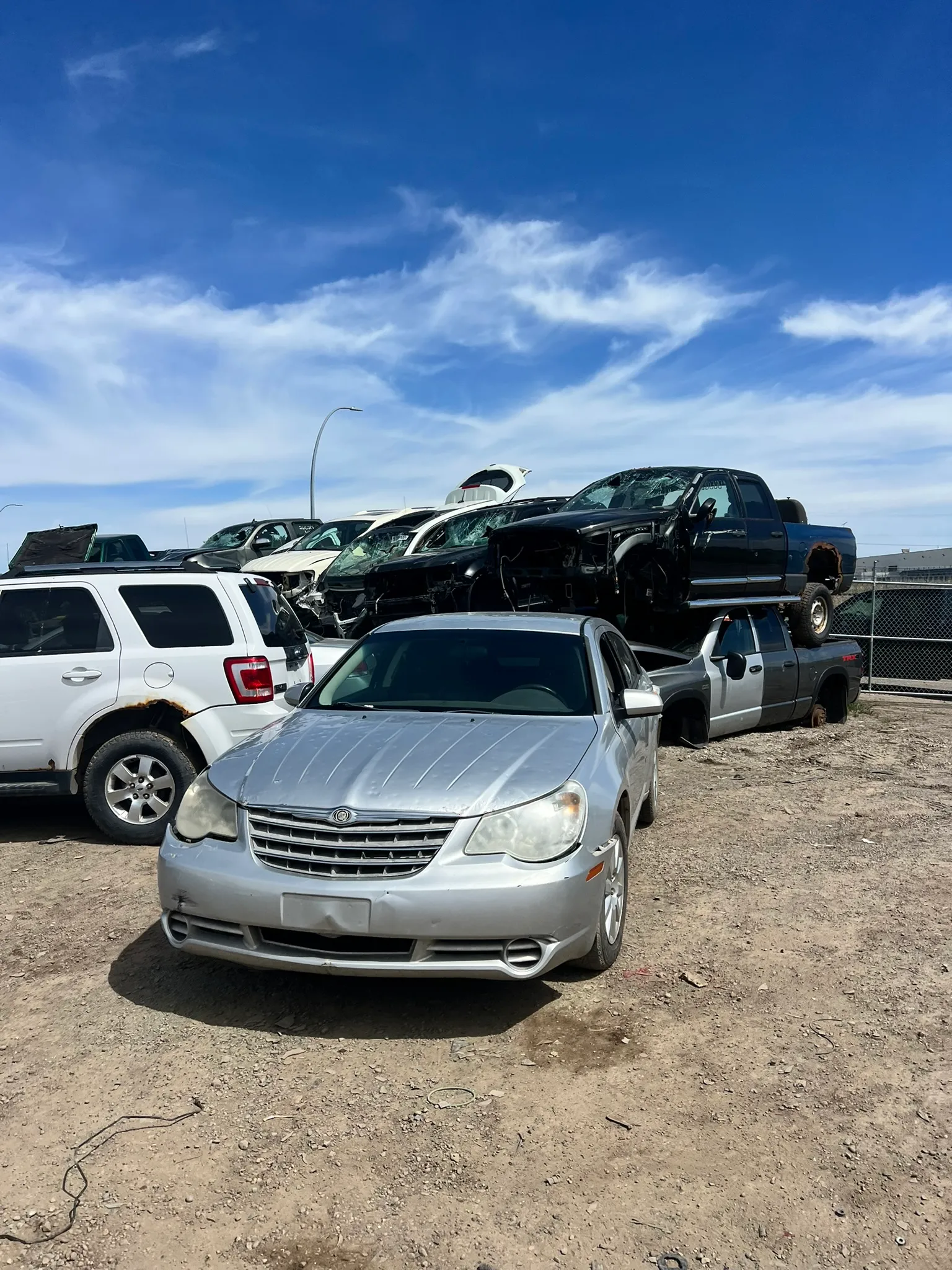 Silver Chrysler sedan with damaged bumper parked in salvage yard; stacked vehicles, chain-link fence, and blue sky with clouds in background.