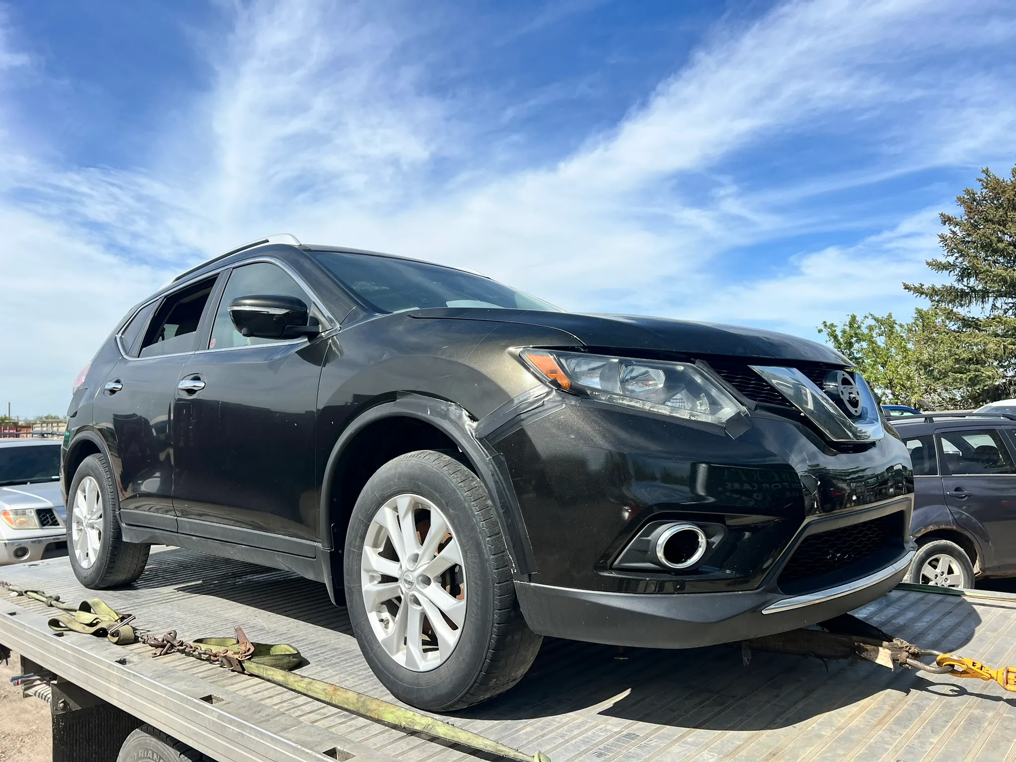 Black Nissan Rogue with minor front fender damage secured on flatbed tow truck; yellow and green straps, trees and parked cars in background.