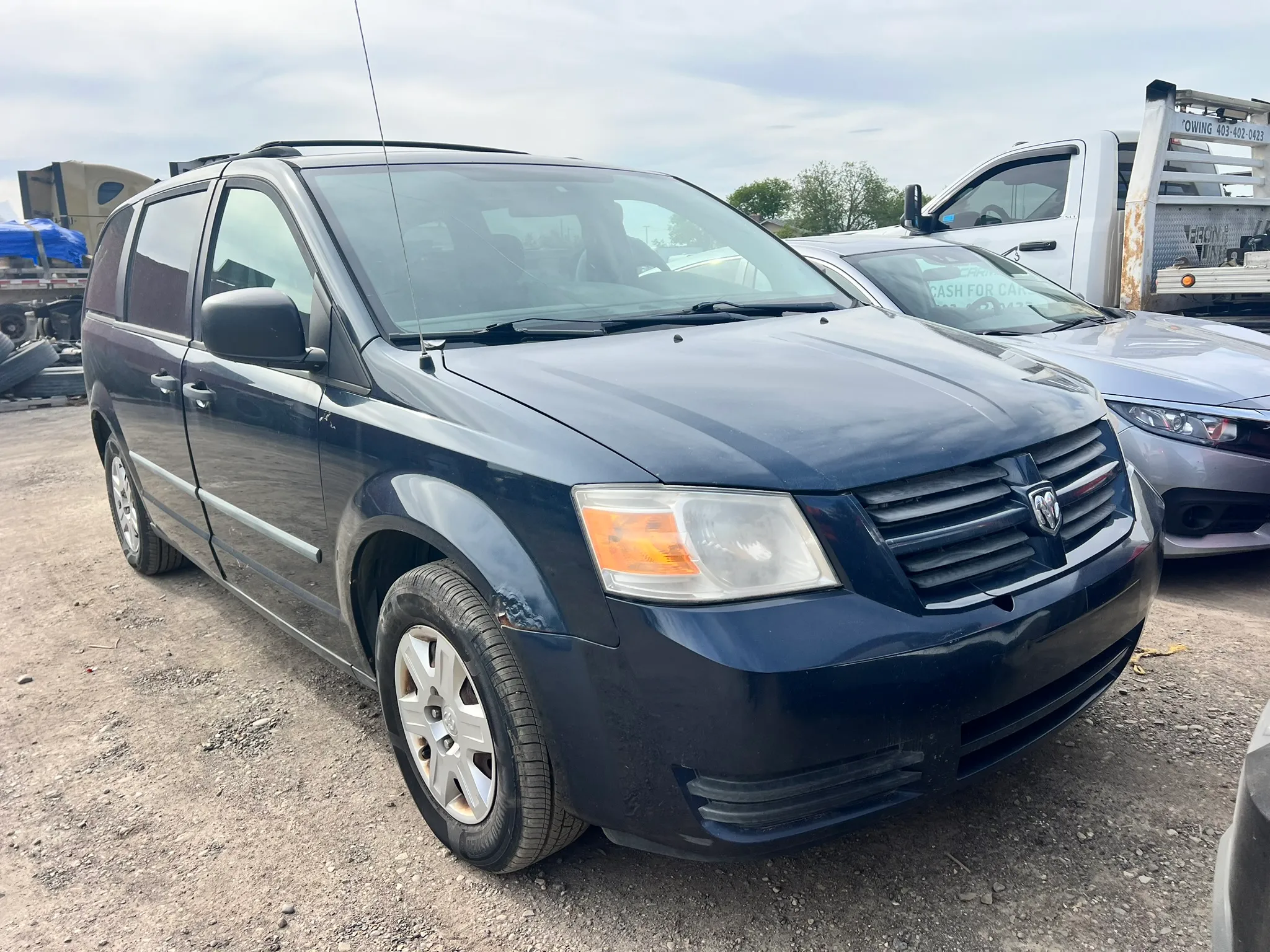 Dark blue Dodge Grand Caravan parked in lot with visible wear; front grille and alloy wheels shown, surrounded by other vehicles.