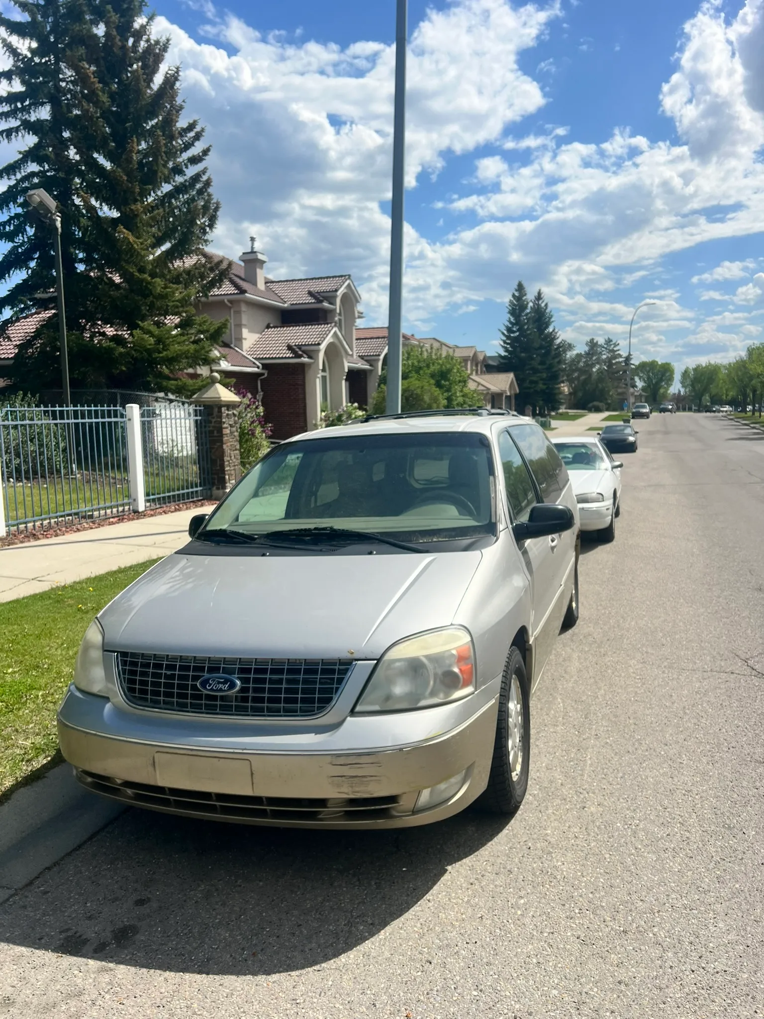 Silver Ford minivan parked on suburban street with other vehicles; white fence, pine tree, and partly cloudy sky in background.