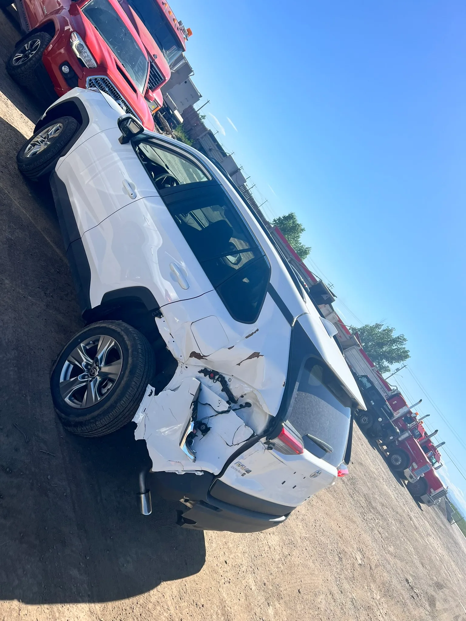 White SUV with crumpled rear side and misaligned wheel parked in lot; red SUV and fire trucks visible under sunny sky.