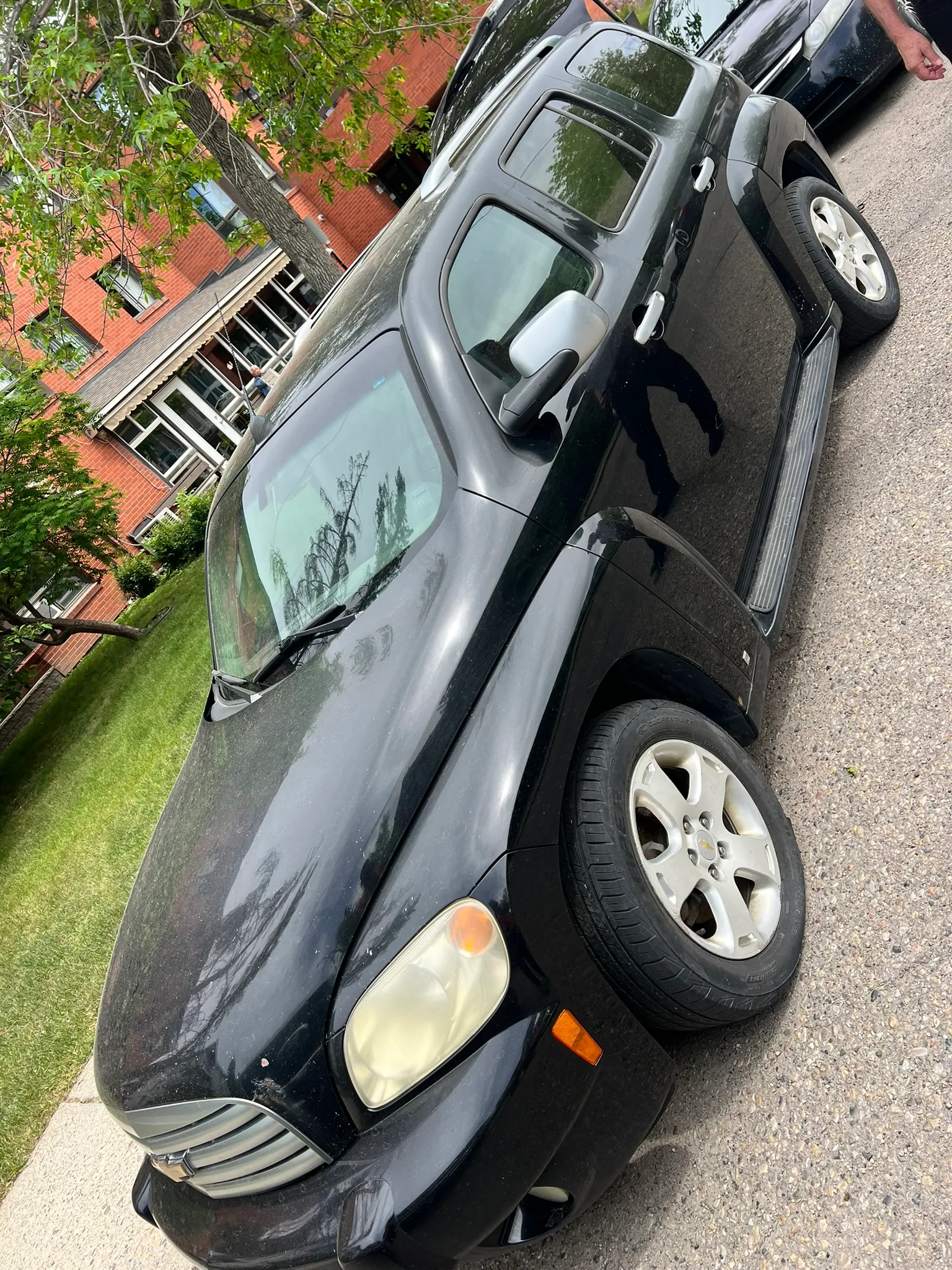 Black Chevrolet HHR parked near grassy area with front wheel turned outward; person standing nearby, red brick building in background.