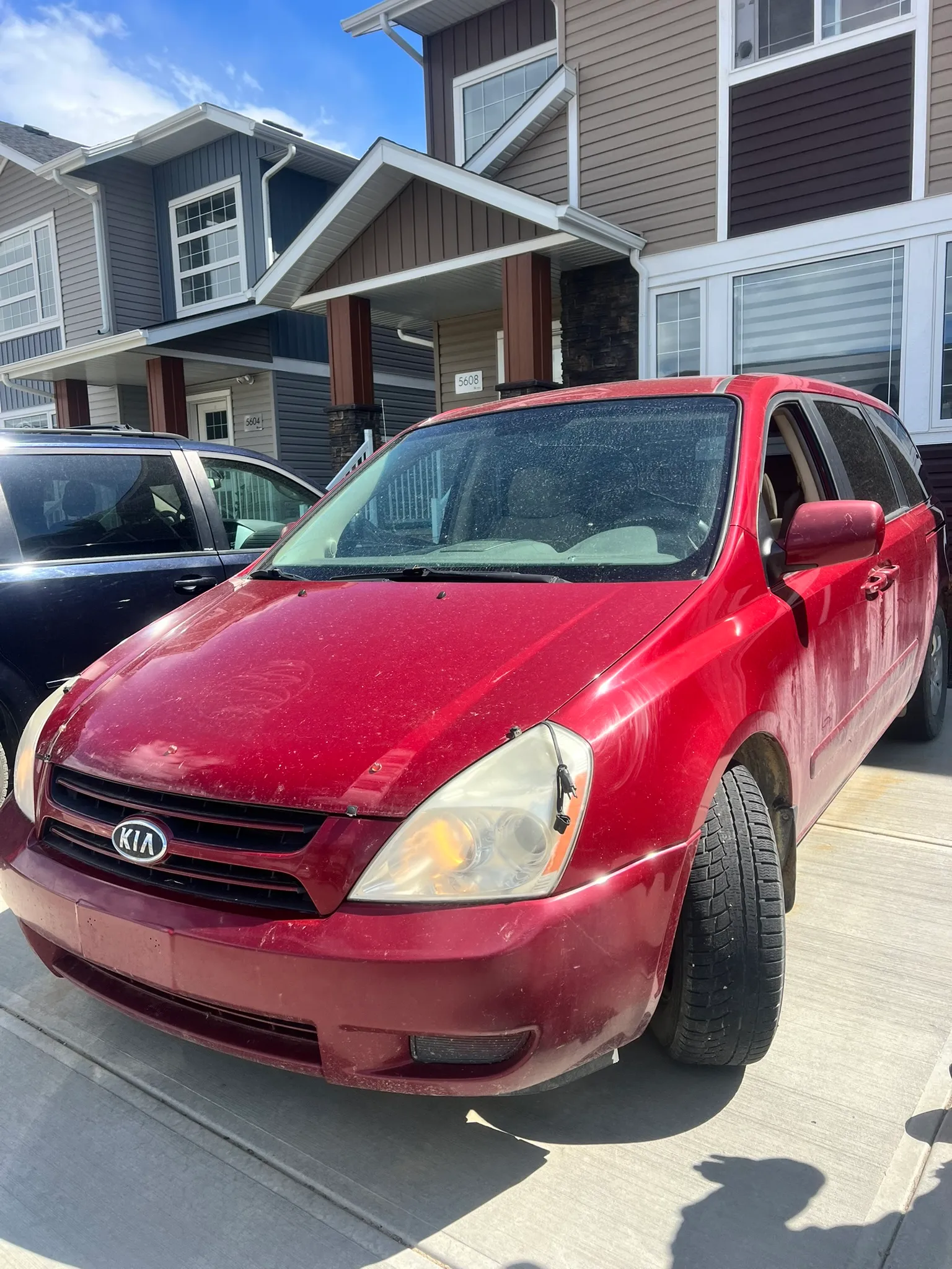 Red Kia minivan with headlights on parked in residential driveway; front wheel turned outward, modern house and clear sky in background.
