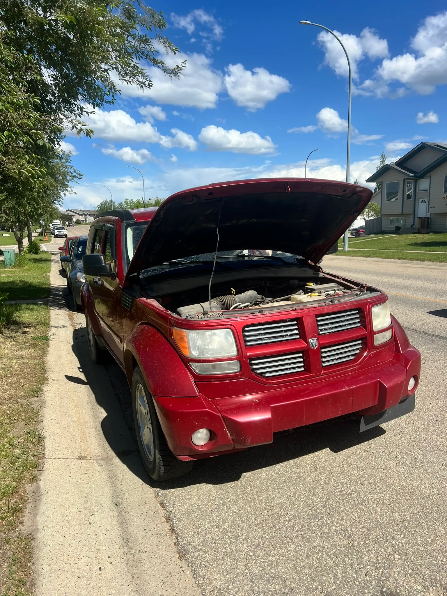 Red Dodge SUV with hood open parked on residential street; engine exposed, sunny day with trees, houses, and scattered clouds.