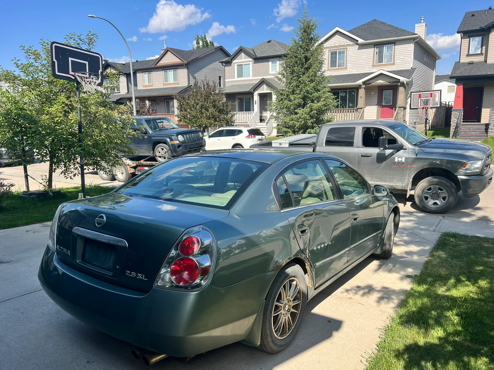 Dark green Nissan Altima with rear-side damage parked in driveway; Dodge Ram truck, basketball hoops, and sunny sky in background.