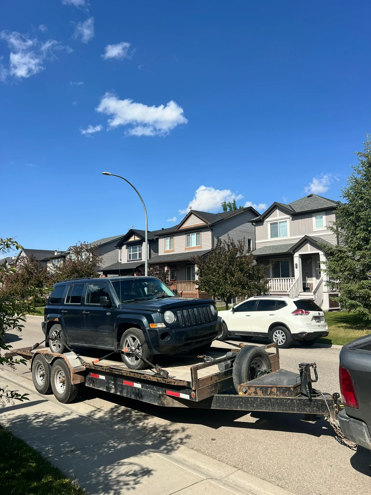 Black Jeep Patriot SUV loaded on dual-axle flatbed trailer; spare tire visible, parked on sunny residential street with modern houses.