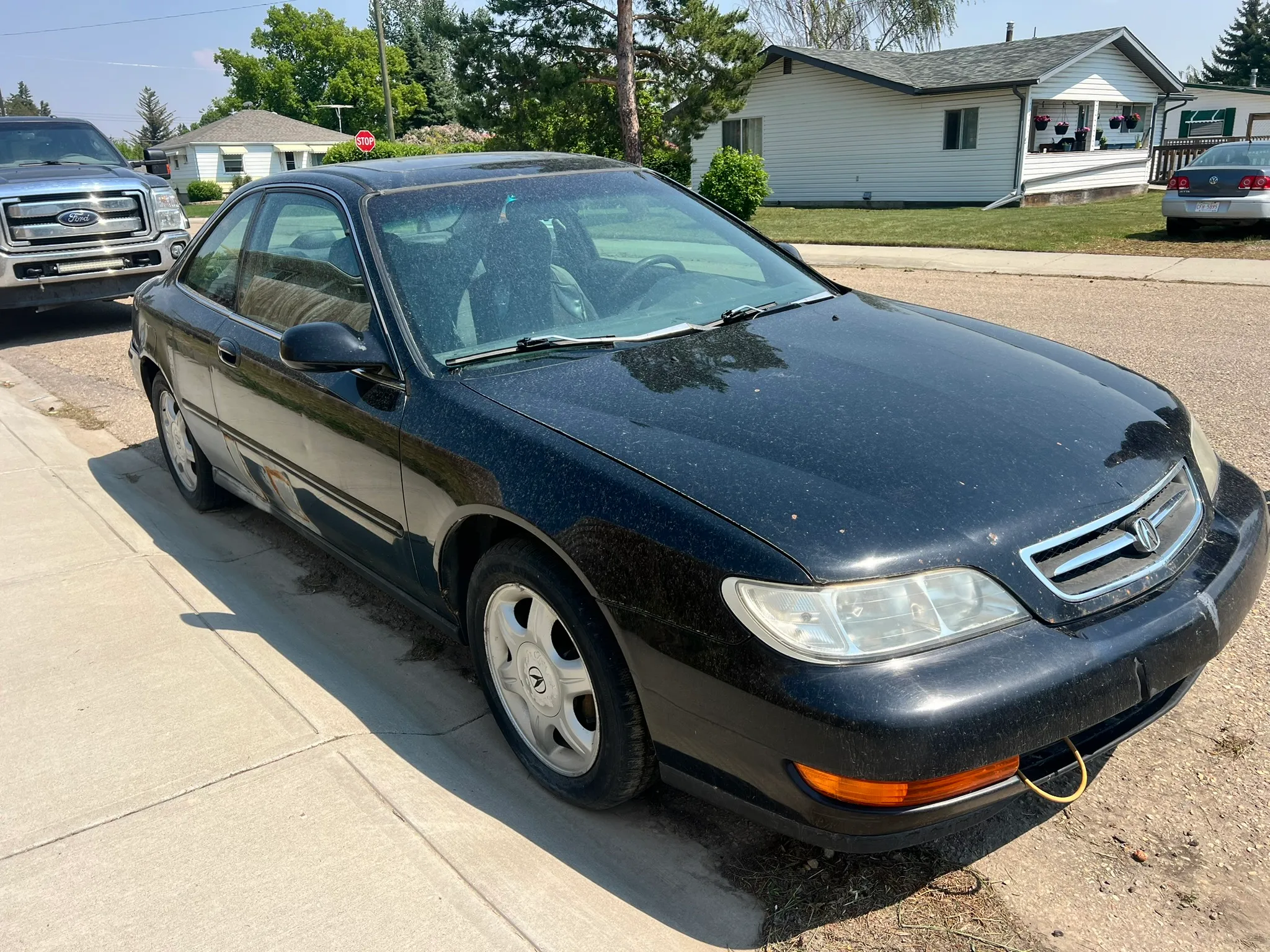 Black Acura CL coupe with yellow cord on front bumper parked on suburban street; dusty exterior and trees in background.