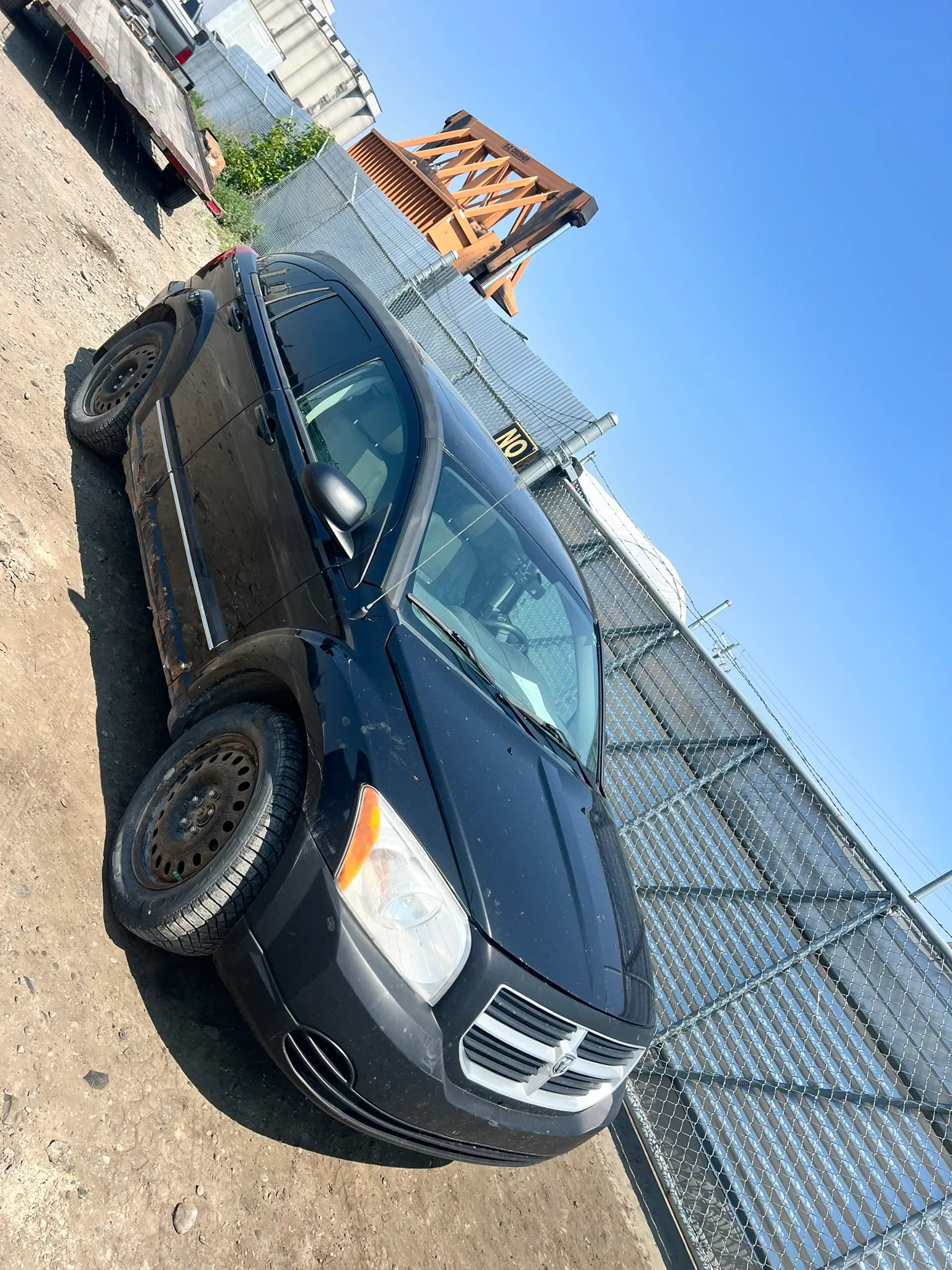 Black Dodge Caliber with spare front tire parked on dirt near barbed-wire fence; industrial structure and trailer in background.
