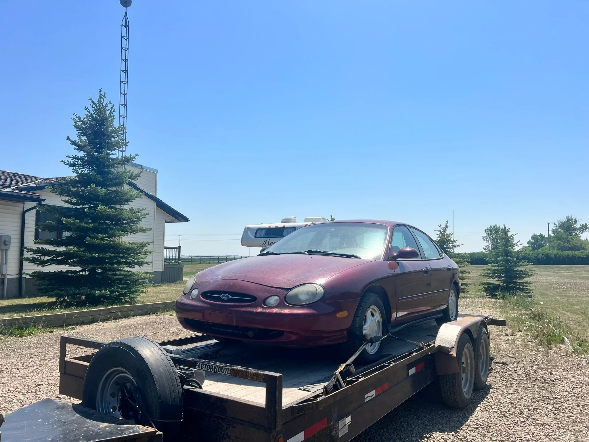 Maroon Ford Taurus sedan loaded on flatbed trailer near white house; gravel lot, evergreen trees, and clear blue sky in background.