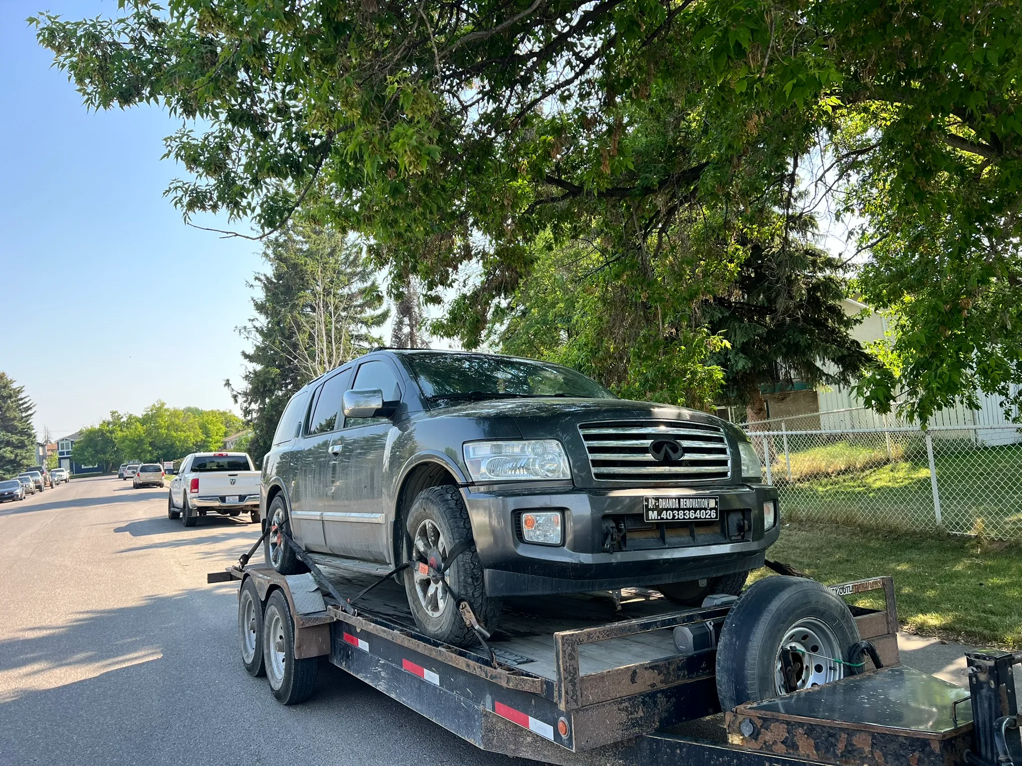 Black Infiniti SUV with dealership plate loaded on flatbed trailer; white pickup truck and shaded residential street in background.