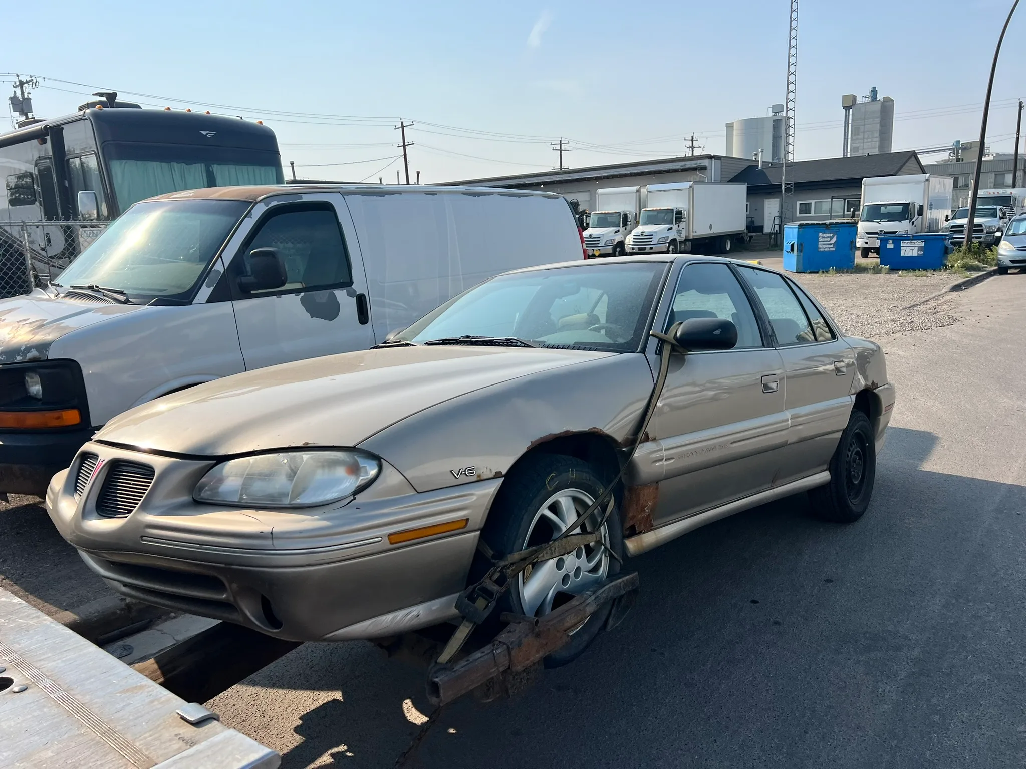 Damaged beige Pontiac Grand Prix missing front wheel, secured for towing near industrial lot with dumpsters and utility poles.
