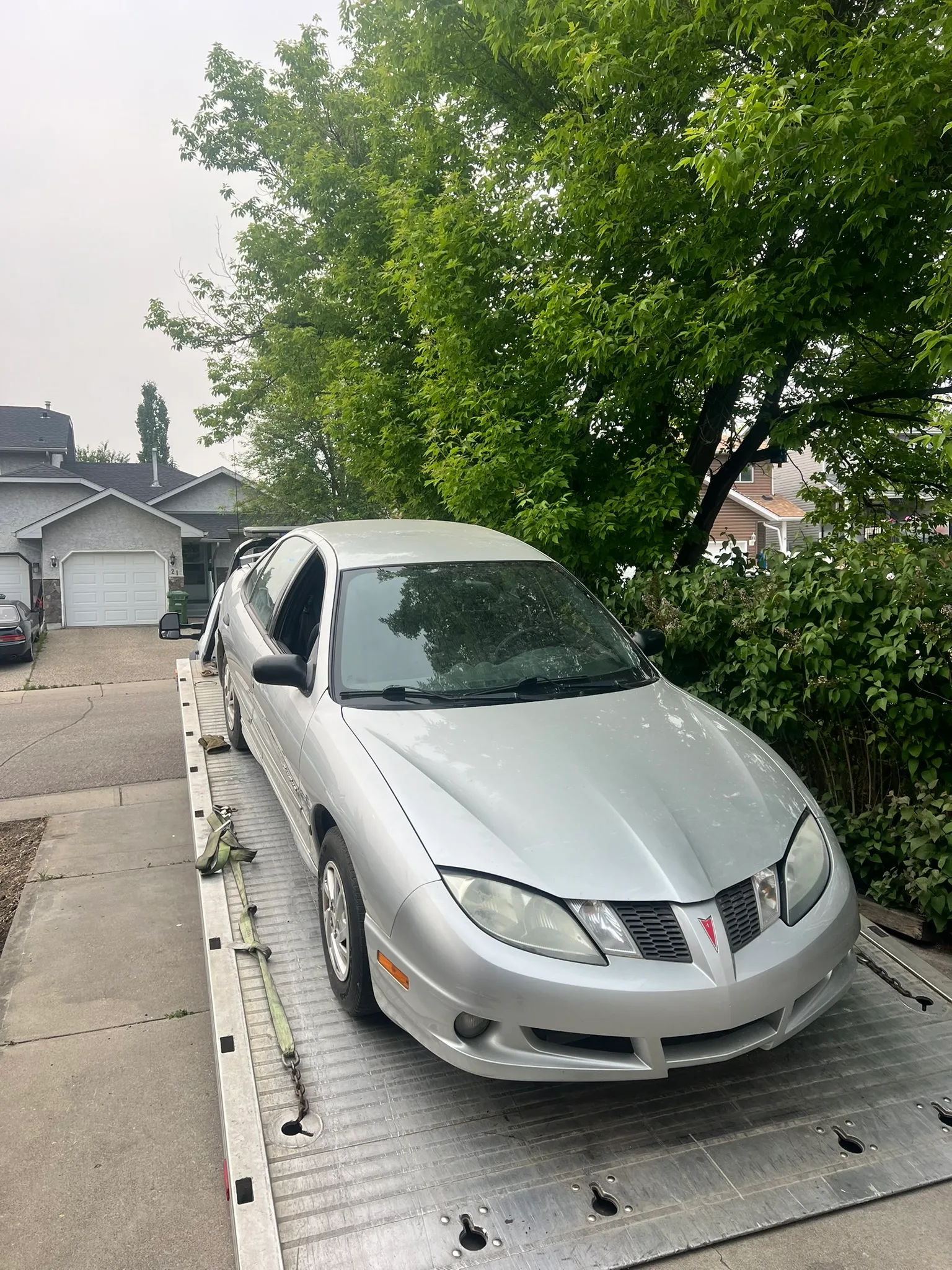 Silver Pontiac sedan secured on flatbed tow truck in residential driveway; overcast sky with trees and houses in background.
