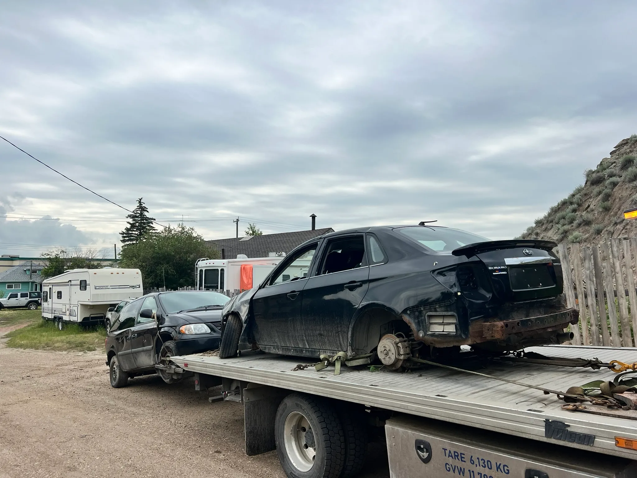 Two damaged black sedans loaded on flatbed tow truck; missing bumper and taillights, gravel road with RV and houses in background.