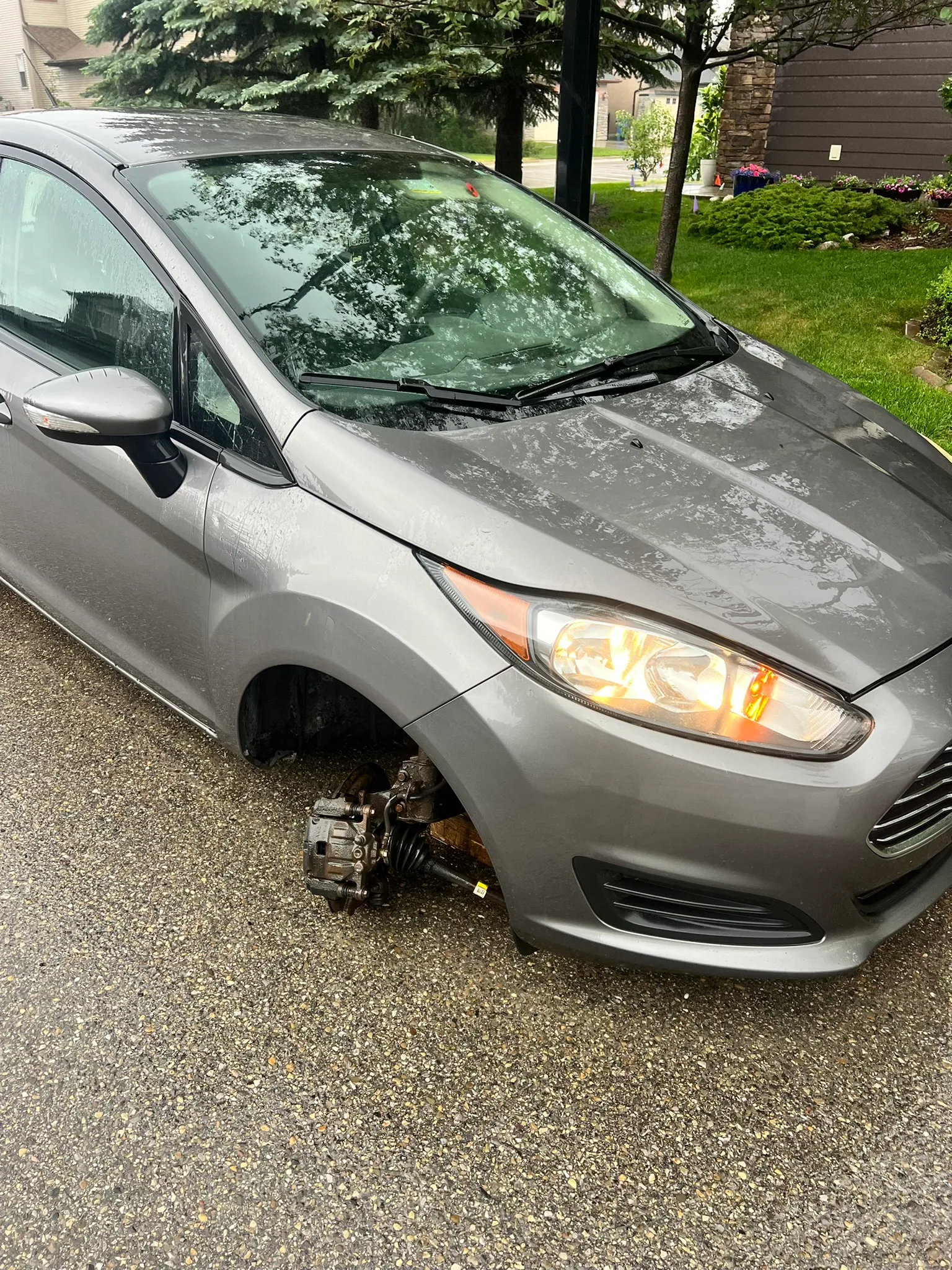 Gray Ford Fiesta parked on wet residential street with missing front left wheel; exposed brake and suspension components visible, headlights on, and green lawns and houses in background.
