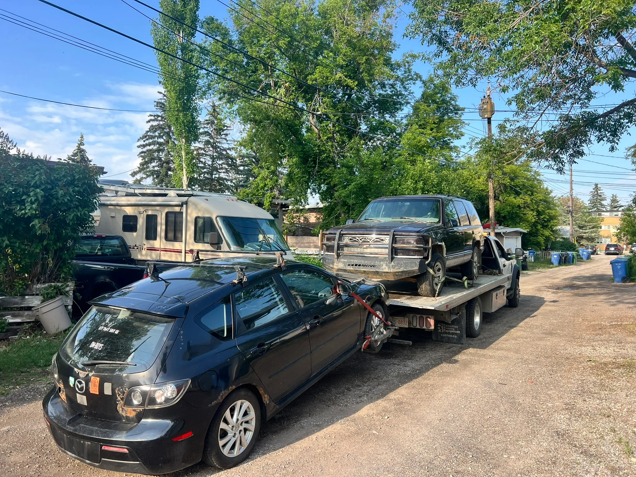 Damaged black Mazda hatchback with bent hood towed behind flatbed carrying SUV; residential alley with RV and garbage bins visible.