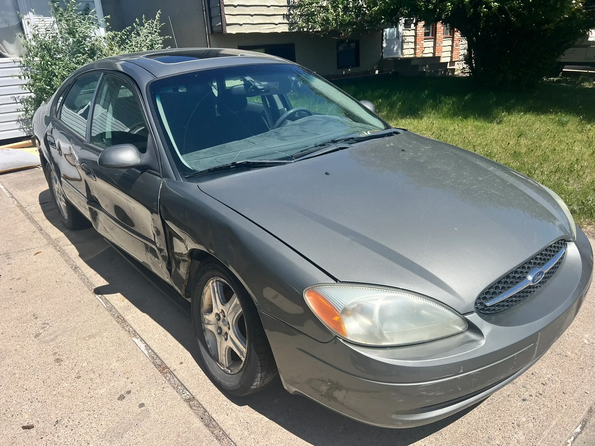Gray Ford sedan with dented driver’s door parked on concrete driveway in Blackie; sunny day with trees and houses in background.