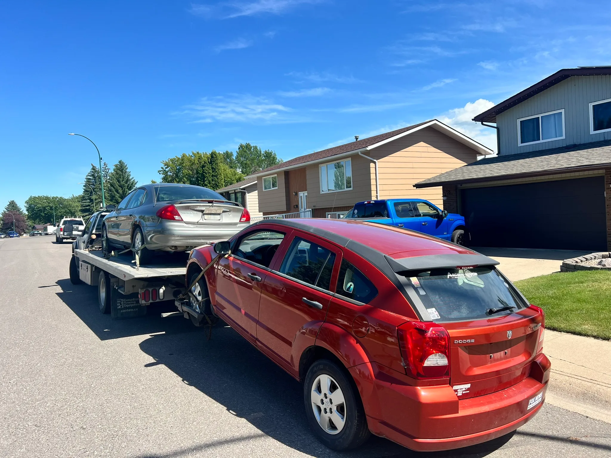 Red Dodge Caliber being towed behind flatbed carrying silver sedan on sunny suburban street in; houses and lawns visible.