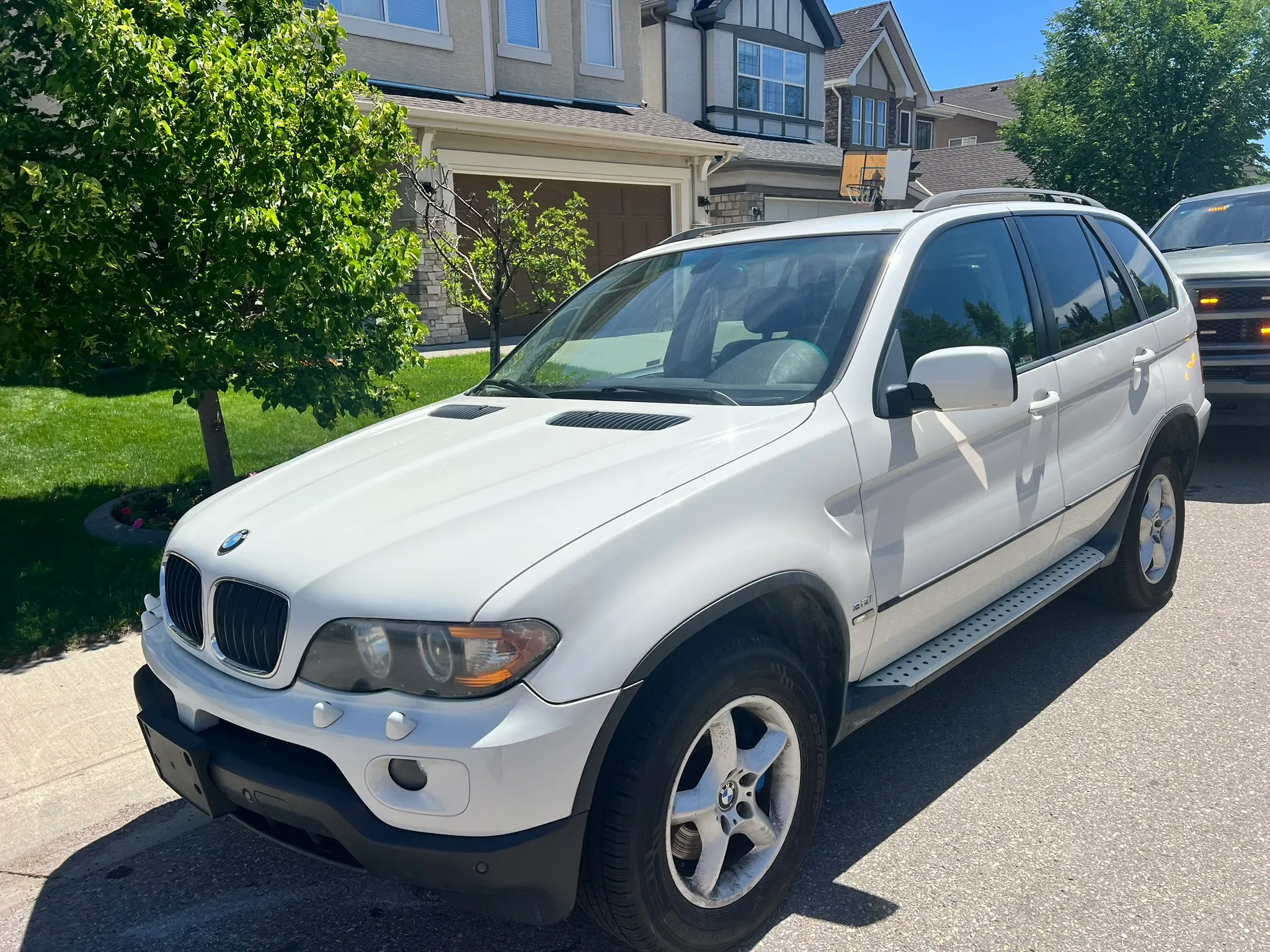 White BMW X5 SUV parked near curb on sunny day; modern homes, tree, and manicured lawn in background.