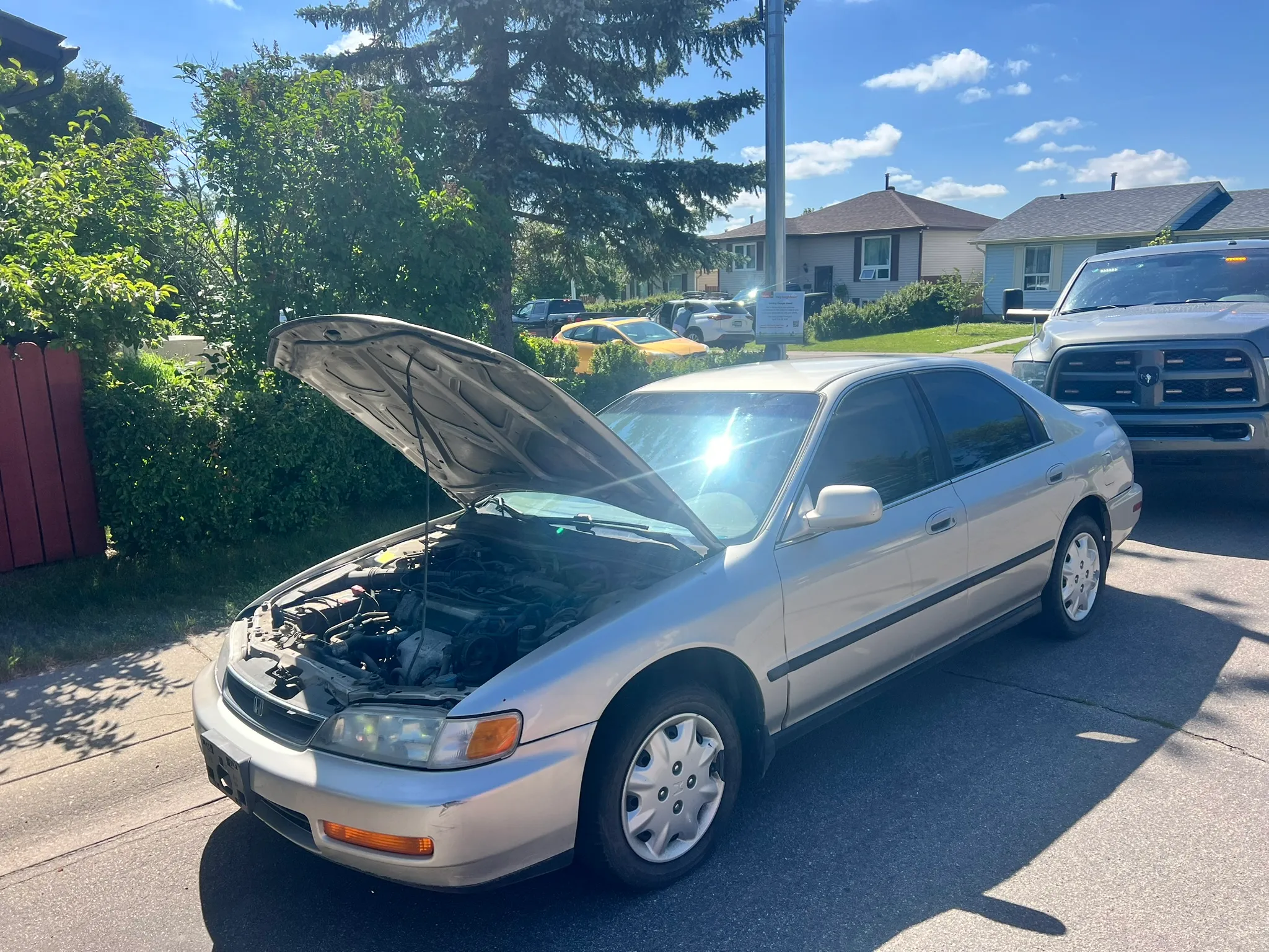 Silver sedan with hood open on a suburban street, surrounded by trees and houses; older model, likely undergoing inspection.