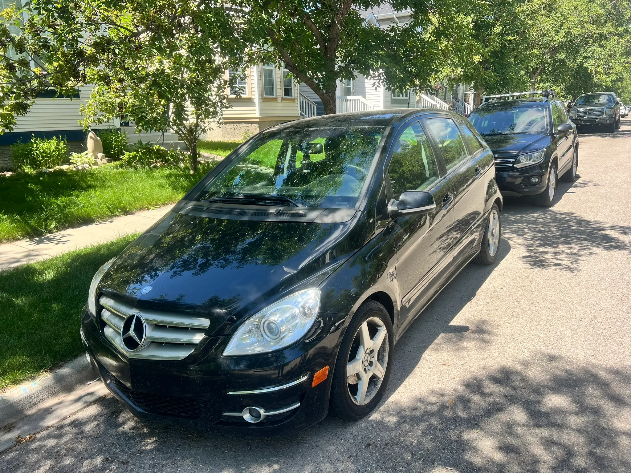 Black Mercedes-Benz B-Class parked on shaded residential street in Blackie; sunny day with trees, lawns, and houses in background.
