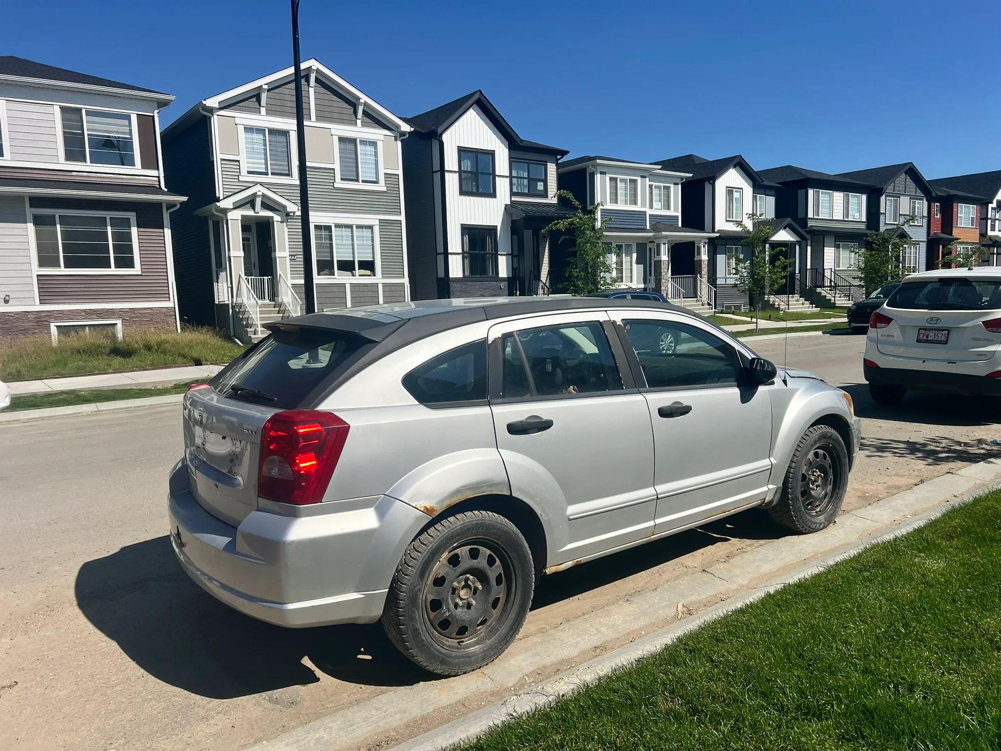 Silver Dodge Caliber with rear wheel rust parked on sunny suburban street in Blackie; modern homes and clear blue sky in background.