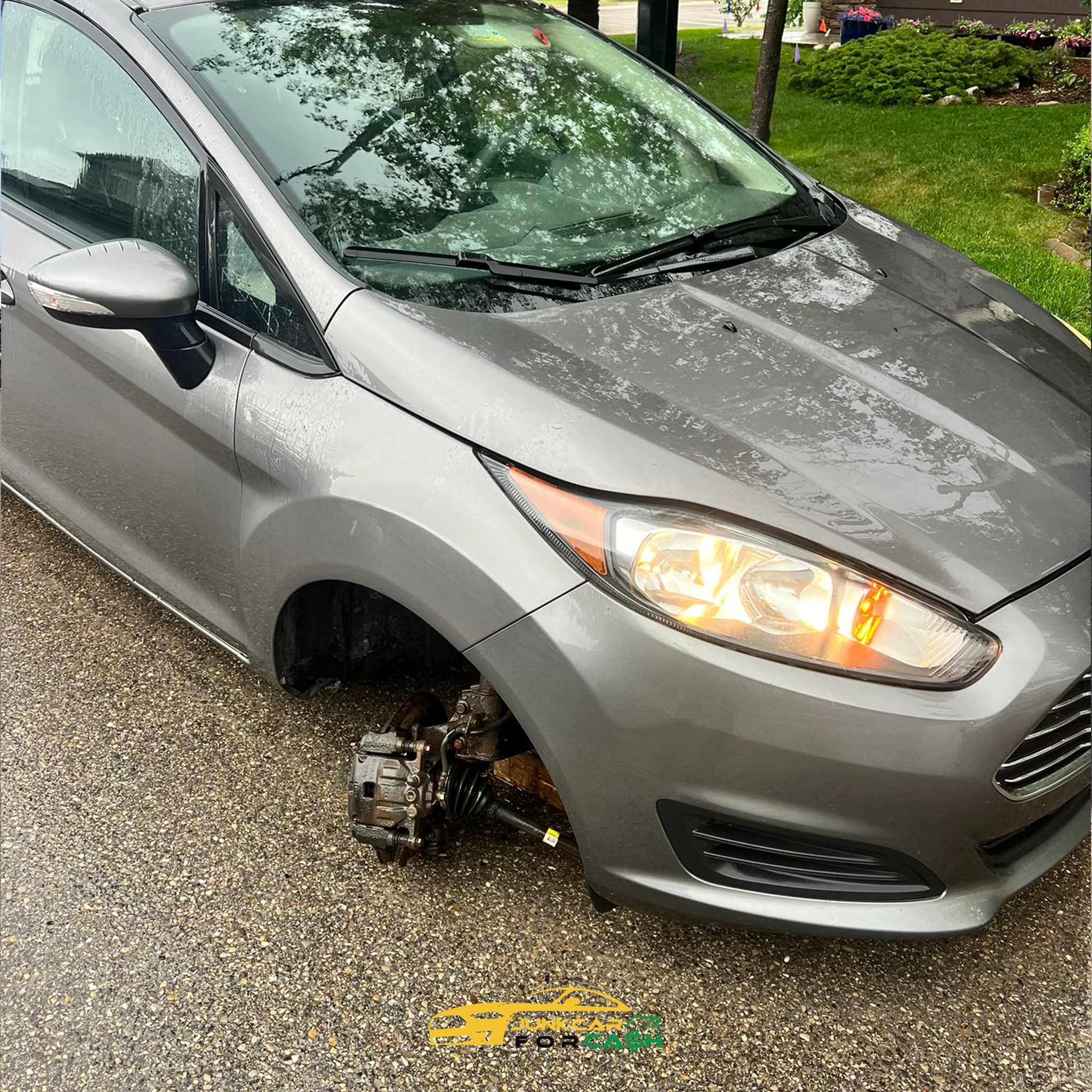 "Gray Ford Fiesta missing front driver's side wheel, resting on brake rotor in wet suburban street after rain."