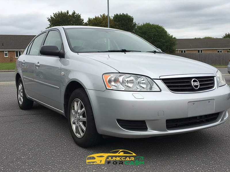 Silver sedan parked on a residential street, shown from the front-left angle with clear headlights, alloy wheels, and houses in the background under an overcast sky.