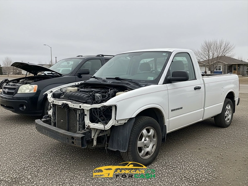 White pickup truck with its front end removed, exposing the engine, parked beside another damaged vehicle in a lot.