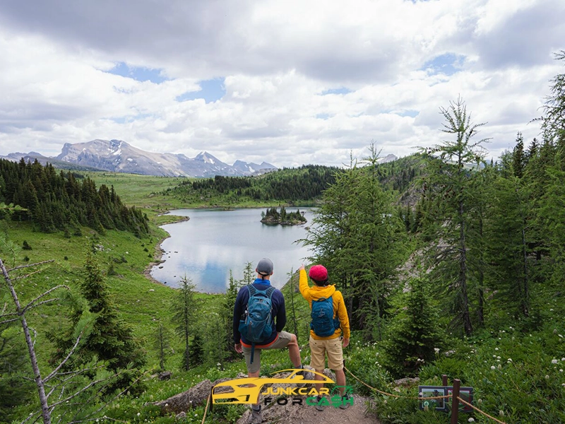 Two hikers with backpacks overlook a scenic mountain lake surrounded by evergreen forest and distant peaks under a cloudy sky.
